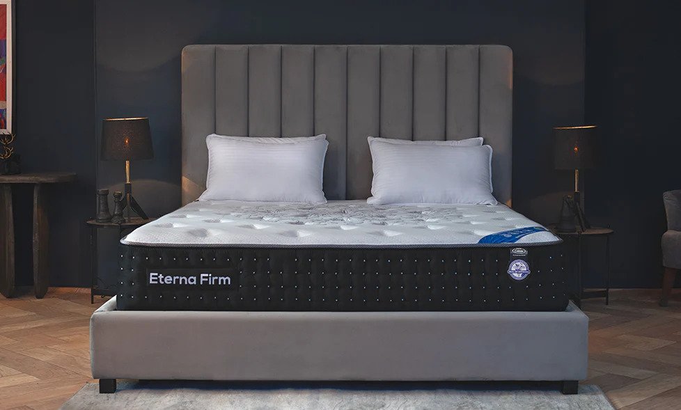Celeste Eterna Firm mattress in a modern bedroom with gray headboard, white bedding, and black side panel featuring tufted design and certification badge.